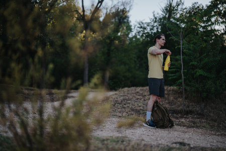 Man prepares water bottle on forest trail during outdoor hike with backpack nearby