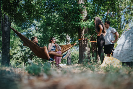 Friends enjoying a relaxed camping day in the forest with a hammock, tents, and casual conversationの写真素材