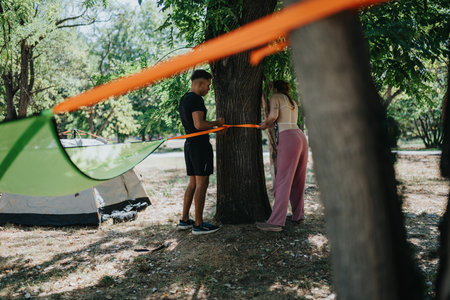 Couple and friend set up an orange strap between trees at a sunny park campsite for outdoor...
