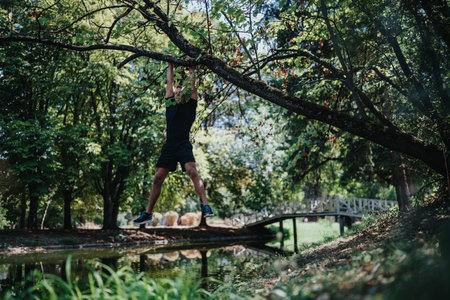 Man hangs from a tree branch during an outdoor workout in a park near a wooden bridge