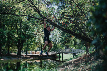Man swings from a tree branch over a pond in a lush park setting, capturing outdoor fitness and adventurous energyの写真素材