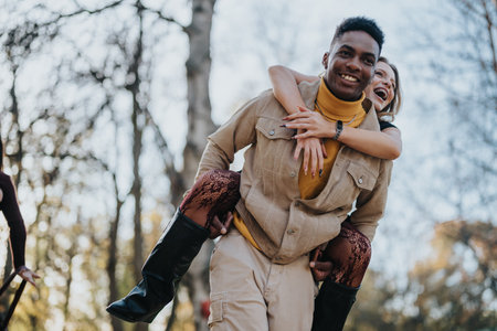 Joyful couple sharing a piggyback ride in a sunny park during autumn, showcasing friendship, romance, and playful energyの写真素材