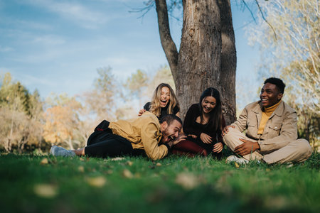 Friends Enjoy a Laughing Moment Together in the Park Under a Large Tree on a Sunny Dayの写真素材