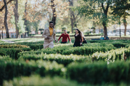 Diverse friends in a sunny park enjoy running, laughter, and togetherness among manicured hedges and autumn lightの写真素材
