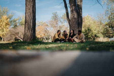 Group of friends sitting on the grass in a park under trees, enjoying conversation and outdoor...