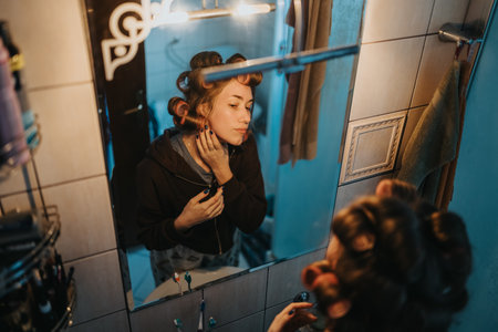 Woman with curlers looks in bathroom mirror during at-home grooming momentの写真素材