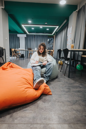 Young woman studies on a bean bag in a modern collaborative workspace, brainstorming and writingの写真素材