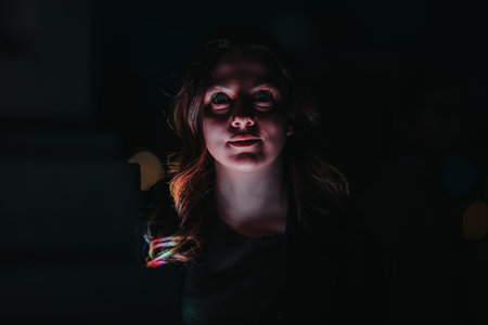 Young woman at night glow from laptop screen with surprised expression in dark city settingの写真素材