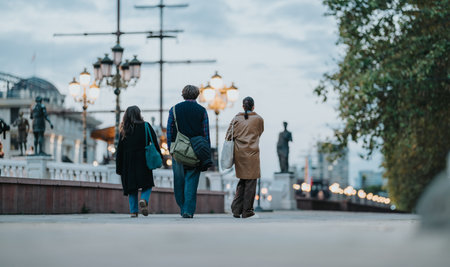 Three friends walk along a city promenade at dusk, carrying bags amid warm streetlamps and urban sceneryの写真素材