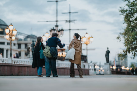 Friends chat and walk along a twilight city riverfront with lampposts, ships, and a warm urban glowの写真素材