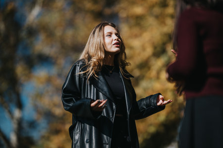 Woman in a leather jacket speaks outdoors in a park during autumn sunlight and warm moodの写真素材