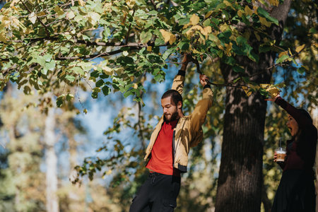 Man hangs from a tree branch in a sunny park while a friend offers popcorn and drinks