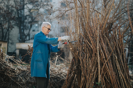 Elderly man in blue coat cuts a bundle of sticks with a handsaw outdoors