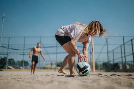 Woman bends to pick up volleyball on sunny beach court during outdoor sportの写真素材