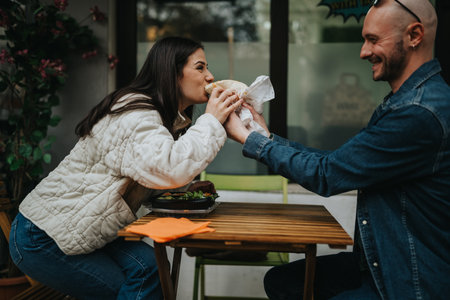 Two friends share a sandwich at an outdoor cafe, smiling and enjoying a casual meal togetherの写真素材