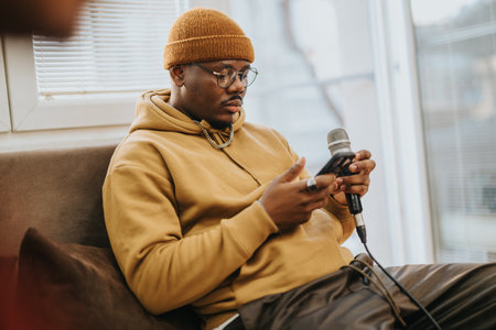 Young man in hoodie and beanie recording a podcast at home with microphone and smartphoneの写真素材