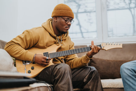 Man in mustard hoodie plays electric guitar on sofa in cozy living room at home