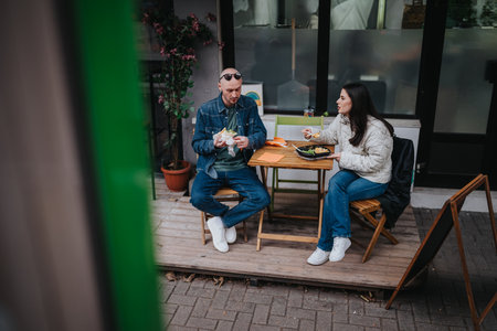 Two friends enjoy a casual outdoor meal at a small cafe table on a wooden deck