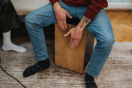 Man sitting on a cajon drum in casual home setting with tattoos and jeansの写真素材
