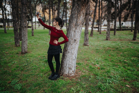 Teen girl taking a selfie in a park, leaning against a tree in a red top and black skirt