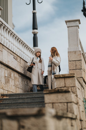 Two women in winter coats converse on stairs in an urban outdoor setting