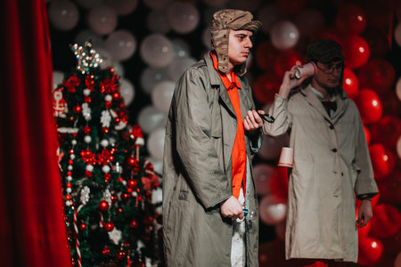 Two men in trench coats on a Christmas theater stage beside a decorated tree and balloons