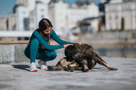 Woman in teal athletic wear pets a large dog on a waterfront promenade with city backdrop and bright dayの写真素材