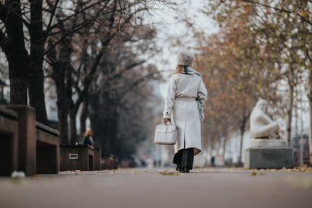 Woman in a light coat walking away in a tree-lined park on a cold winter day, with a bag in handの写真素材