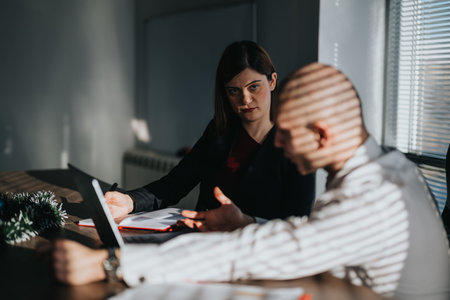 Two business employees discuss documents during a daytime office meeting by a window