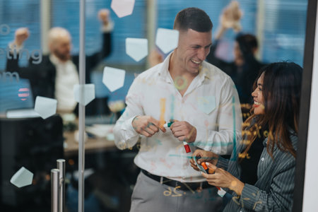 Two professionals laugh and brainstorm at a glass-walled office with sticky notes and markers