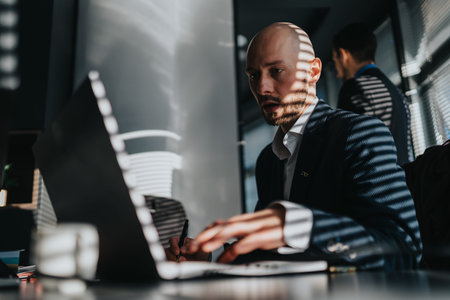 Focused business man works on laptop at a modern office desk with striped light shadows