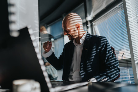 Confident businessman celebrates at his desk in a modern office with striped sunlight patterns