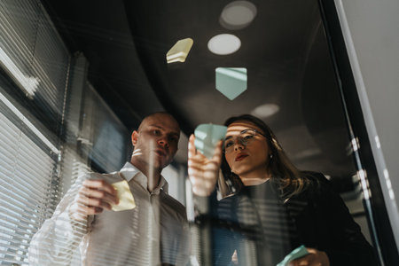 Two professionals collaborate in a glass-walled office, placing sticky notes and sharing ideas