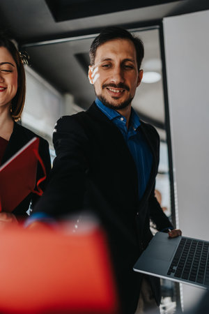 Confident businessman extends hand toward camera with laptop in a modern office, coworker beside himの写真素材