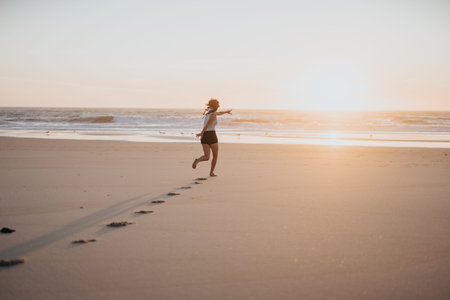 Girl runs along the sunset beach at golden hour, leaving footprints in the warm sandの写真素材