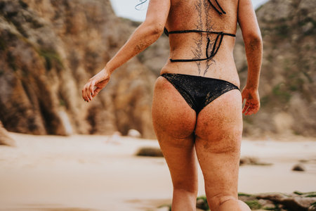Woman in black bikini walking on sandy beach with tattoos visible on back and rugged rocky backdrop