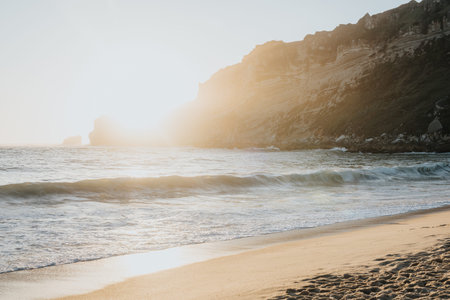Person sitting on a sunny beach at sunset near rocky cliffs and gentle wavesの写真素材