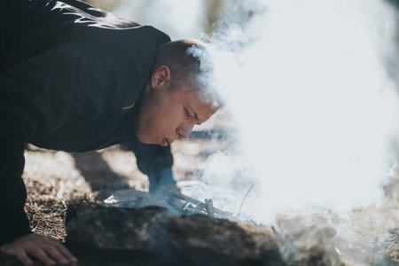 Man crouching by campfire with rising smoke outdoors, focused on the flame