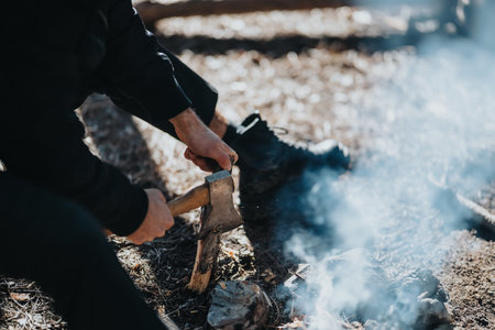 Person chopping wood with an axe beside a campfire outdoors, wearing dark clothing and bootsの写真素材