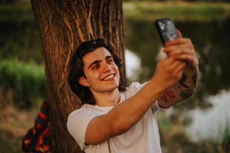 Young man taking a selfie outdoors, smiling under a tree by a lake during sunset