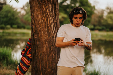 Young man using smartphone outdoors by a lake under a tree in a park setting