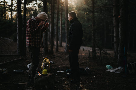 Two men camping in a pine forest at dusk with gear and a stove nearbyの写真素材