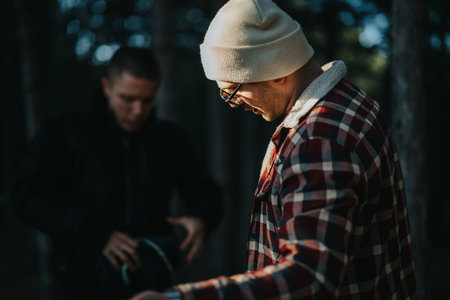 Man in plaid jacket and beanie outdoors with friend in a forest setting during duskの写真素材