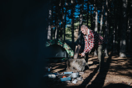 Man chopping wood at a camping site with a tent and campfire setup in the woods