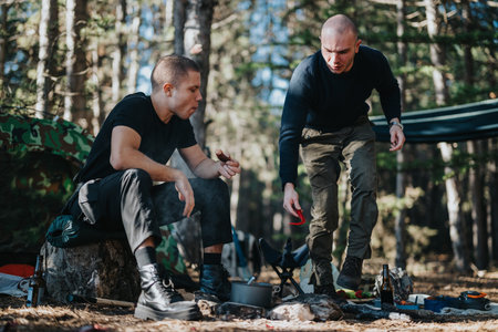 Two men camping in a forest campsite cooking over a fire with gear and tent nearby