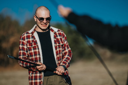 Smiling man in plaid jacket and sunglasses holds a pole outdoors in a sunny nature setting