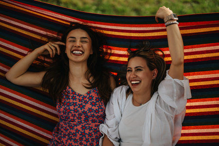 Two friends laugh together under a colorful striped hammock in a sunny outdoor settingの写真素材