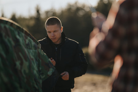 Man in a black jacket helps with gear near camouflage tent as friends photograph outdoors