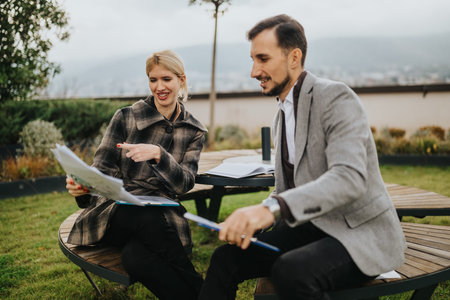 Business partners discuss documents outdoors on a circular wooden bench, smiling and collaborating