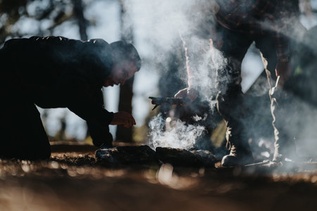 Crouching figure tending a campfire outdoors with smoke and friends nearby